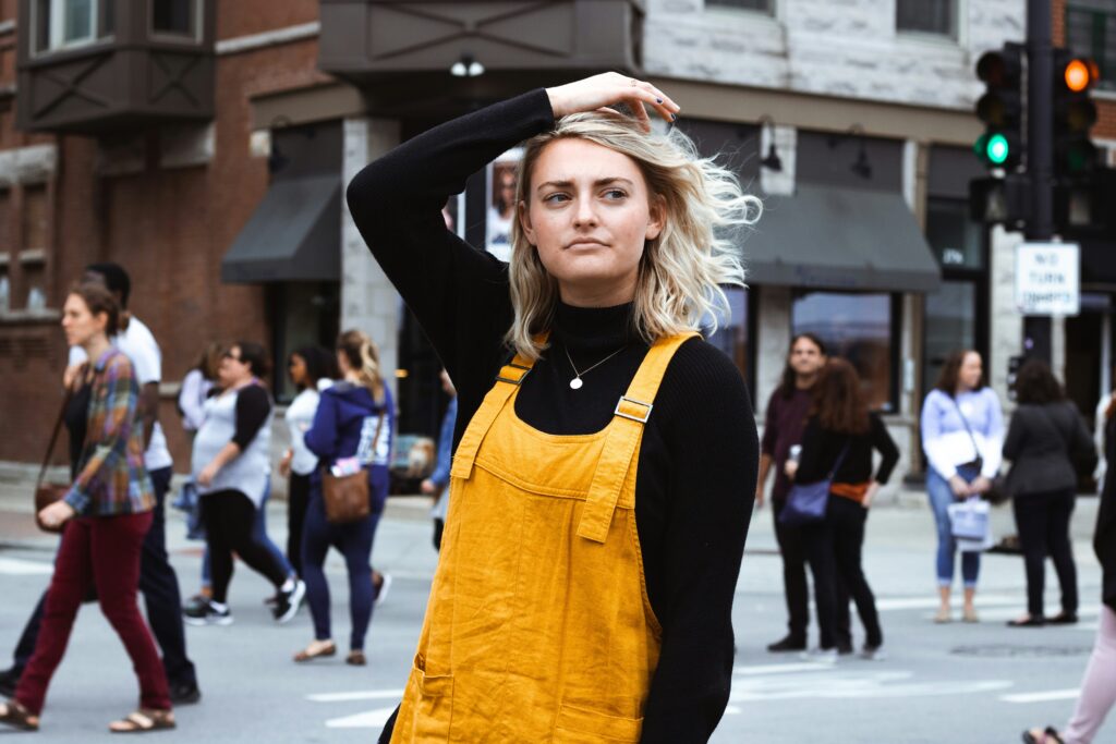 A woman with blonde hair crossing a city street looking thoughtful and wearing yellow dungarees and a black poloneck.